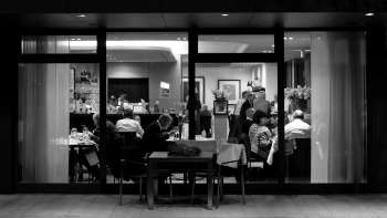 Black and white photo of a busy restaurant with a view from outside through the glass front.