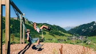 Two children swinging on a mountain playground with a view over the summer panorama of Malbun in Liechtenstein