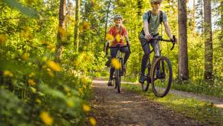 Two cyclists with helmets ride relaxed on their gravel bikes on a sunny forest path through a green canopy - active vacation in Liechtenstein's nature