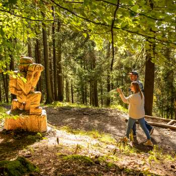 Woman points to a detailed wooden sculpture in the forest on the Walser Saga Trail
