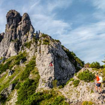 Hikers on a rocky mountain path in front of a rugged ridge with a red and white signpost