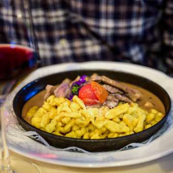 Hearty spaetzle with beef ragout, garnished with tomato, served in a pan - accompanied by a glass of red wine on a laid table.