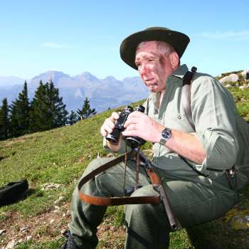 A hunter with binoculars sits in a meadow against an alpine mountain backdrop.
