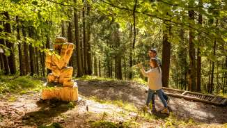 Two visitors discover a carved wooden figure along the WalserSagenWeg in Triesenberg, Liechtenstein - mystical themed hiking trail in the forest