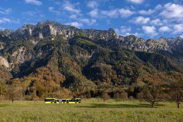 A yellow and black LIEmobile bus drives through an autumnal meadow landscape in front of high mountains