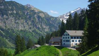 Traditional alpine and mountain inn Sücka in Liechtenstein, nestled in a green mountain landscape with views of wooded slopes and snow-capped peaks. Traditional alpine and mountain inn Sücka in Liechtenstein, nestled in a green mountain landscape with views of wooded slopes and snow-capped peaks.