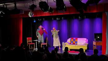 A cabaret scene at K-BUM in Liechtenstein—a colorful stage with a sofa, a duo of actors, and an audience in the hall