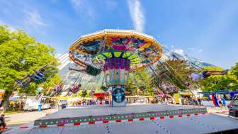 Large colorful chain carousel at the fair in Balzers, full of happy visitors in front of a mountain backdrop under a blue sky.