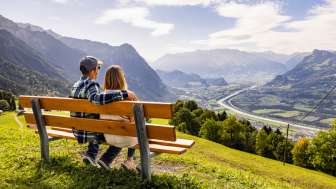 Couple sitting on a bench in Triesenberg looking out over the Rhine Valley in Liechtenstein with an impressive Alpine panorama