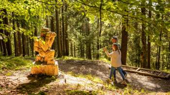 Two hikers walk past a wooden figure on the Walser Saga Trail