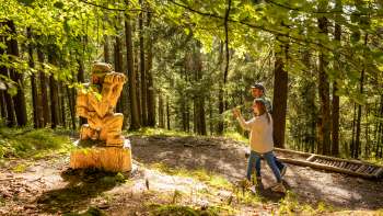 Two hikers walk past a wooden figure on the Walser Saga Trail