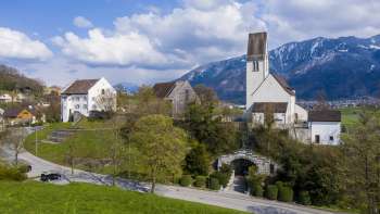 Panoramic view of the "Kirchhügel" (church on the hill)  Bendern with its historic church, surrounded by well-tended paths, trees and traditional architecture against an Alpine mountain backdrop.