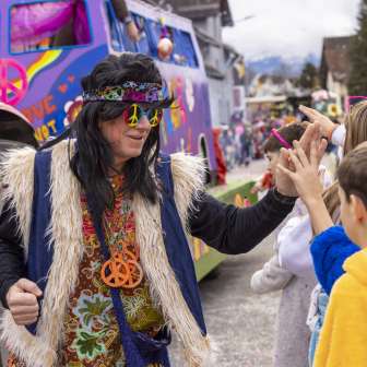 Hippie dressed up at the children's carnival in Mauren, high-fives with children in front of a colorful float.