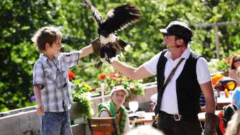A boy holds a bird of prey in his arms, guided by a falconer