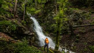 Hikers with rucksacks in front of an idyllic waterfall near Nendeln, embedded in dense forest and moss-covered rocks.