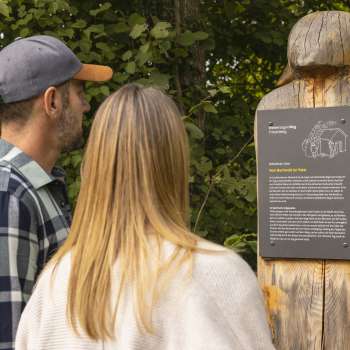 Two people read an information board with a legend on the Walser Saga Trail, on a carved wooden figure