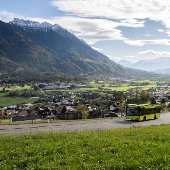 Green LIEmobil bus drives along a winding road above Liechtenstein with a view of the Rhine Valley and the surrounding Alps