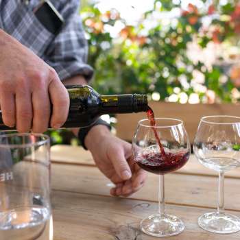 A glass of red wine is poured from a bottle into a wine glass, with flowering plants in the background.