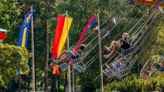 Children and adults ride on a chain carousel in the sunshine at the fair in Balzers, surrounded by colorful flags.