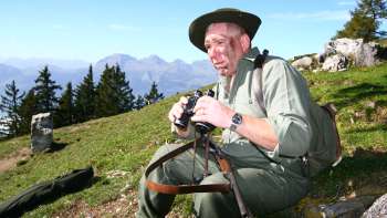 A hunter with binoculars sits in a meadow against an alpine mountain backdrop.