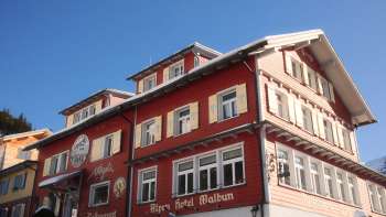 Exterior view of the Alpenhotel Vögeli Malbun with the Elchbar on the first floor - a traditional red building with white lettering against a blue sky.