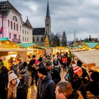 Atmospheric evening shot of the Christmas market in Vaduz 2018 with festively decorated market stalls and crowds in front of the government building and cathedral