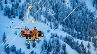 Tina Weirather and Marco Büchel sit together on a chairlift, surrounded by snow-covered trees and the winter landscape in Malbun.