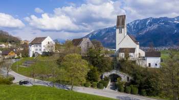 Panoramic view of the "Kirchhügel" (church on the hill)  Bendern with its historic church, surrounded by well-tended paths, trees and traditional architecture against an Alpine mountain backdrop.