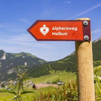 Red sign with the inscription "Alpine Heart Trail Malbun" in front of a picturesque mountain backdrop and summer meadow in Liechtenstein.