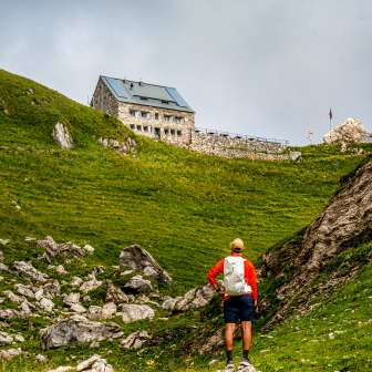 Hiker approaches the Pfälzer hut on a green slope in Liechtenstein, framed by rocks and meadows