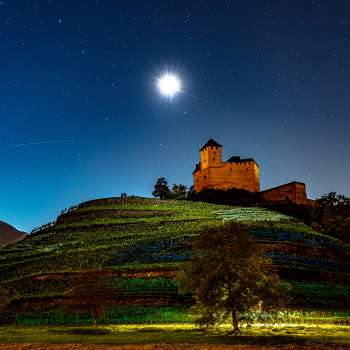 The illuminated Gutenberg Castle at night under a full moon, nestled in the vineyards.