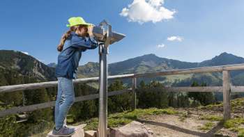 Girl with a green hat looks through a telescope on the Malbun Explorer Trail with panoramic views of the Liechtenstein Alps.
