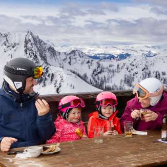 Family enjoying a break on a sun terrace in Malbun with a view of snow-covered mountains.
