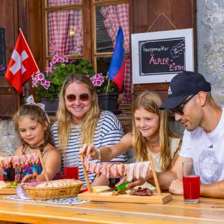 Family sitting on a sunny alpine pasture in Liechtenstein and enjoying a traditional snack with regional specialties and a mountain panorama