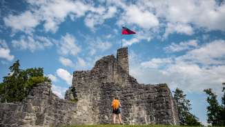 Hiker in front of the historic ruins of Schellenberg Castle with a waving Liechtenstein flag under a blue sky