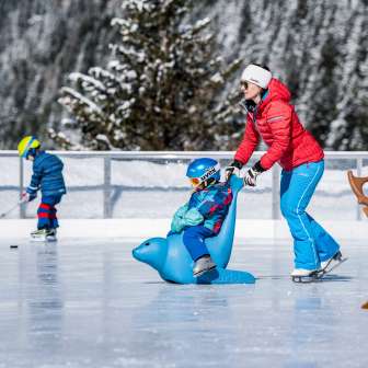Mother pushes child on an ice skating penguin across the ice rink in Malbun, in the background a child with an ice hockey stick