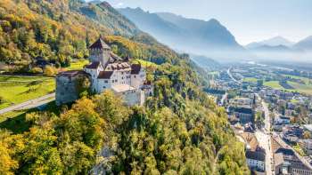 Aerial view of Vaduz Castle on a hill above the city, with a panoramic view of the Alps and the Rhine Valley on a clear day.