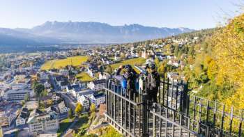Hikers at the railing of the Känzile viewpoint with a panoramic view of Vaduz and the surrounding mountains.
