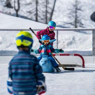 Children play with helmets and field hockey sticks on the ice rink in Malbun, a child sits on a skating helper in front of a goal