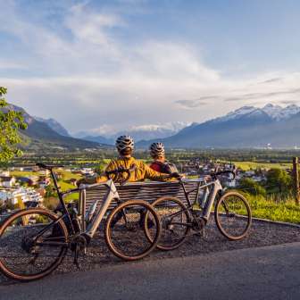 Two cyclists wearing helmets sit on a bench above the Rhine Valley and enjoy the sunset with a view of the Alps - cycling tour highlight in Liechtenstein