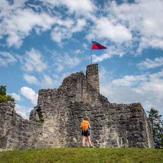 Hiker in front of the historic ruins of Schellenberg Castle with a waving Liechtenstein flag under a blue sky