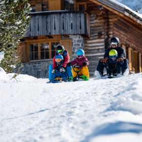 Two adults and three children sit on sledges and start a descent together in front of a snow-covered mountain hut in Malbun