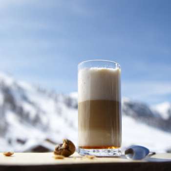 Glass of latte macchiato on a wooden platter against the backdrop of snow-covered mountains.