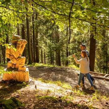 Woman points to a detailed wooden sculpture in the forest on the Walser Saga Trail