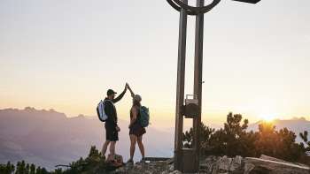 Two hikers give each other a high-five at the summit cross on the Alpspitz at sunrise with a mountain panorama in the background.