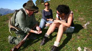 A hunter kneels in front of two laughing women and shows something in his hand.