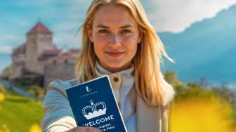 Welcome Adventure Pass - Discover Vaduz & surroundings / AI-generated Woman holding up the WELCOME Adventure Pass in front of Vaduz Castle in a flowering field