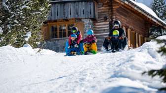 Tobogganing with children in Malbun Two adults and three children sit on sledges and start a descent together in front of a snow-covered mountain hut in Malbun