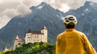 Cyclist with helmet looks at the imposing Gutenberg Castle against a dramatic mountain backdrop in Balzers - cultural tour by bike in Liechtenstein