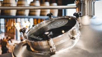 Close-up of a round viewing window on a shiny brewing kettle with a view of the brewing hall in the background.